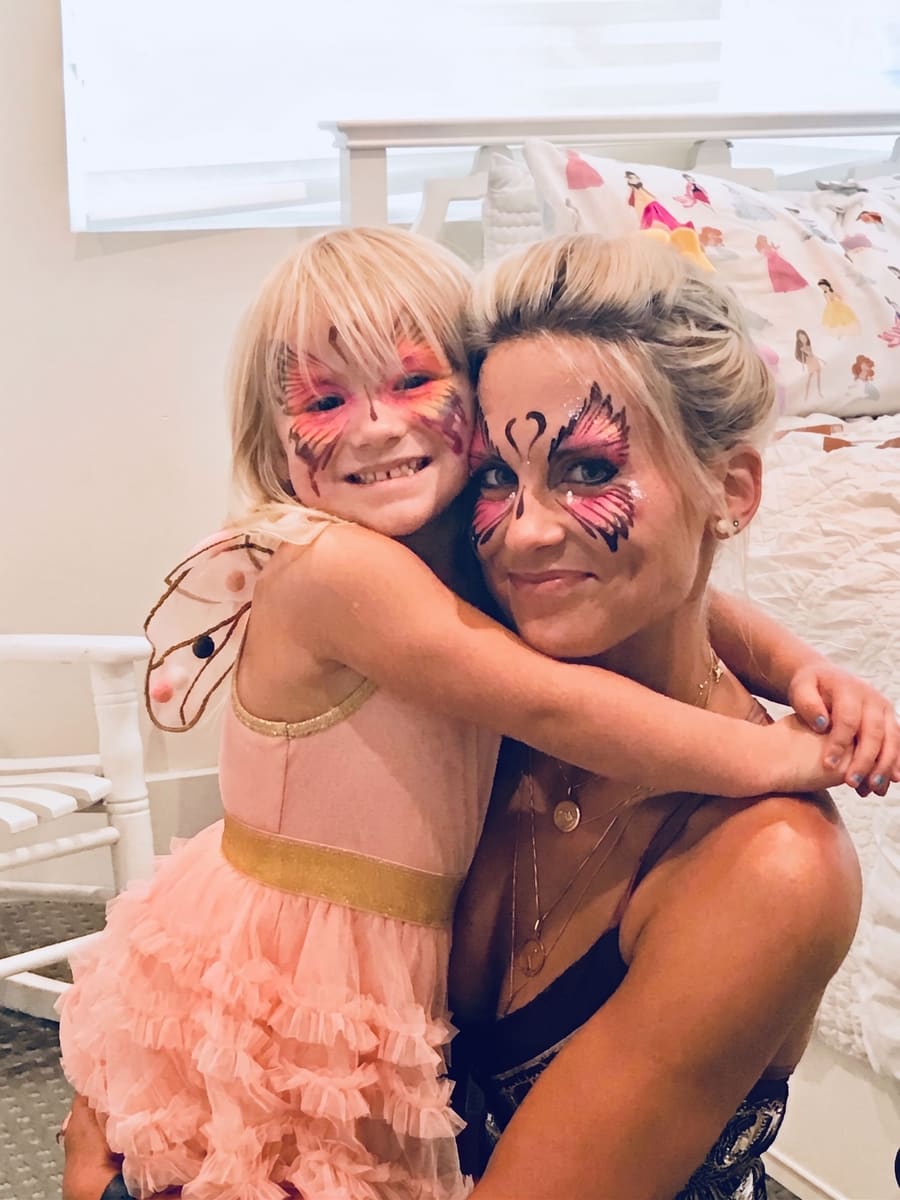 Elisabeth and a little girl in a fairy costume hugging, both wearing pink butterfly face paint